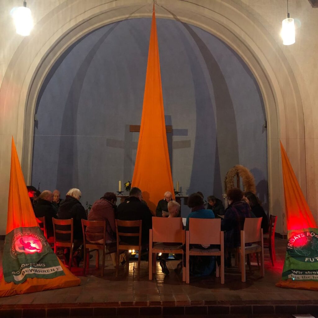 Im Altarraum einer Kirche hängen orange Tücher sowie die Flaggen der Fridays und Christians for Future. Etwa 20 Personen sitzen in einem Stuhlkreis.